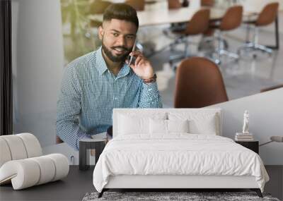 Side view of Indian businessman having a cup of coffee and using internet talking on the phone in modern office Wall mural